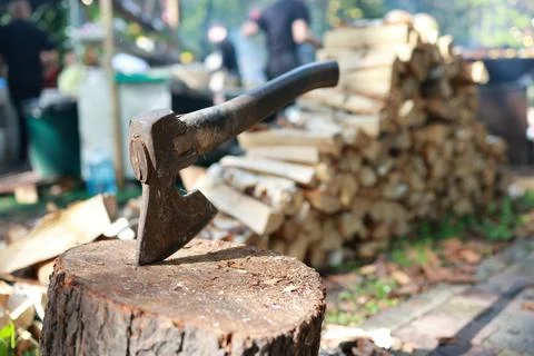 An axe is seen resting on a log in a backyard. Stacks of firewood are organ.. Stock Photos
