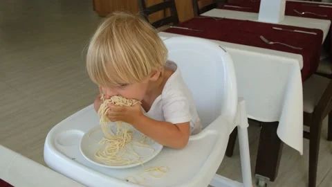 Baby boy eats spaghetti sitting in a baby chair in restaurant Stock Footage 194683239
