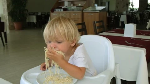 Baby boy eats spaghetti sitting in a baby chair in restaurant Stock Footage 194777130