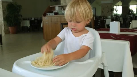 Baby boy eats spaghetti sitting in a baby chair in restaurant Stock Footage 194777168
