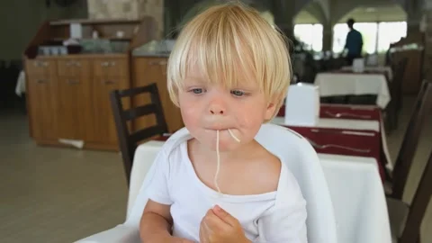 Baby boy eats spaghetti sitting in a baby chair in restaurant Stock Footage 194777217