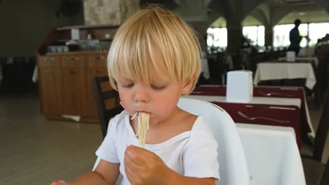 Baby boy eats spaghetti sitting in a baby chair in restaurant Stock Footage 194777249