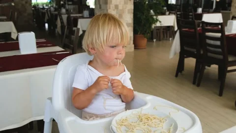 Baby boy eats spaghetti sitting in a baby chair in restaurant Stock Footage 194777526