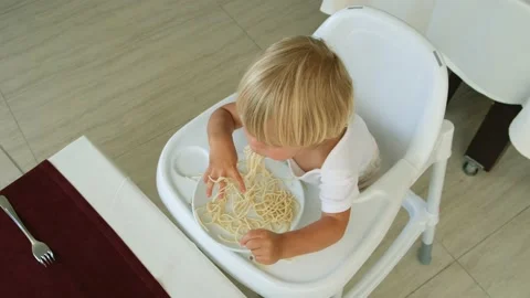 Baby boy eats spaghetti sitting in a baby chair in restaurant Stock Footage 194777611