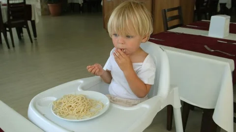 Baby boy eats spaghetti sitting in a baby chair in restaurant Stock Footage 194777707