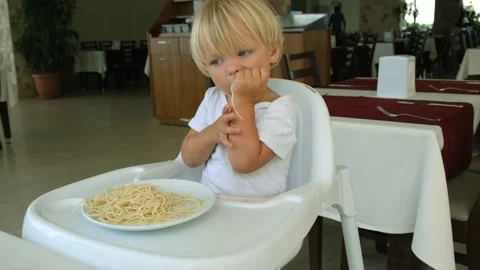 Baby boy eats spaghetti sitting in a baby chair in restaurant Stock Footage 194778021