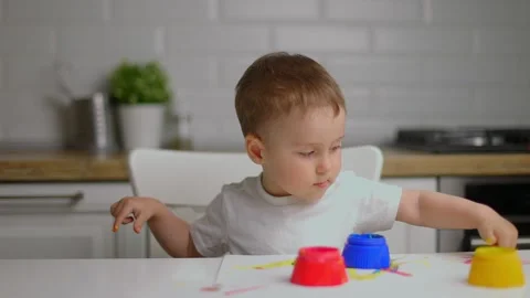 Baby boy sitting behind table and drawing with fingers covered in colorufl paint Stock Footage 156479343
