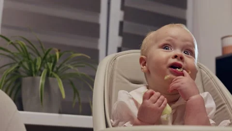 Baby eating solid food in high chair, messy face and hands Vidéo 342504448