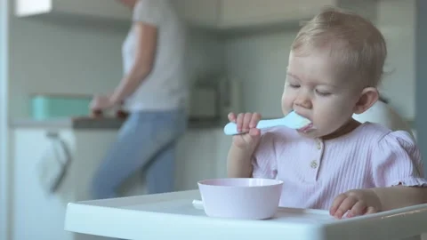 The baby eats with a spoon while mom cooks in the kitchen. Stock Footage 169275815