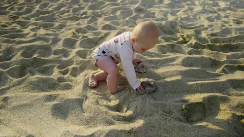 Baby exploring sandy beach, playing with sand, wearing light-colored outfit Stock Footage 313947918