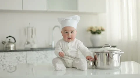 Baby in hat sitting on kitchen table. He holding a soup ladle Stock Footage 23740708