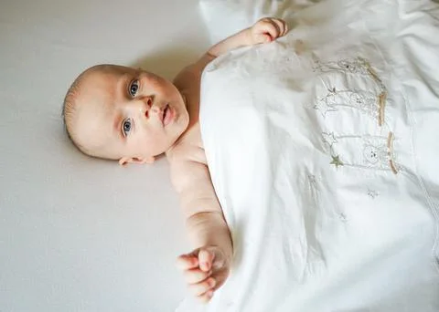 Baby lies on a white sheet under a white blanket Fotos de archivo