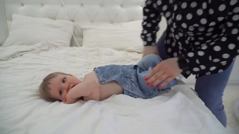 Baby lying on white bed while adult adjusts denim overalls, capturing a tender m Stock Footage 320567720