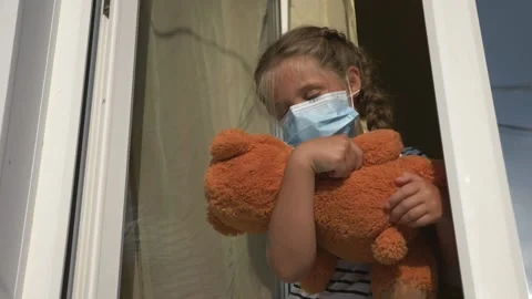 The baby is playing with a Teddy bear in a protective mask and looks out the Stock Footage 137660401