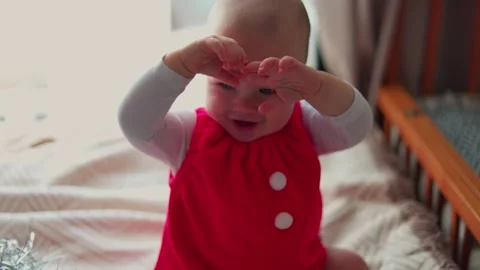 Baby rubs eyes while sitting on a bed in a bright bedroom wearing red festive Stock Footage 347686894