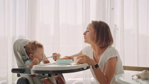 Baby sits at a children's little table. Mum feeds the child with porridge Видео 158509727