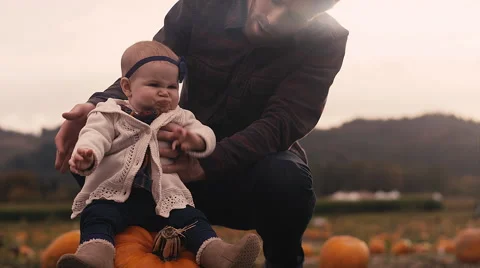A baby sitting on a pumpkin at a pumpkin patch, with her dad holding her up Stock Footage 58593681
