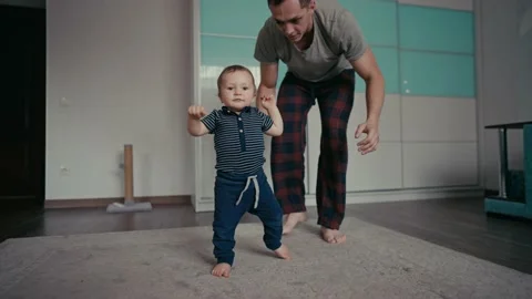 Baby taking first steps with father's help at home. Father Encouraging Baby Son Stockbeeldmateriaal 166604125