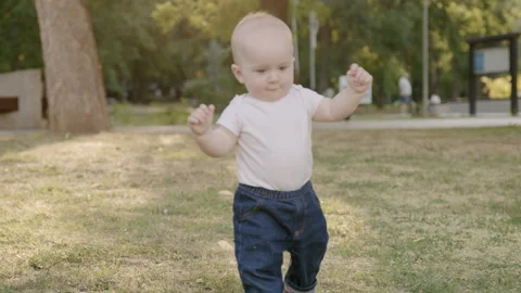 Baby taking first steps in grassy park, holding adults hand, showcasing Stock Footage 315182848