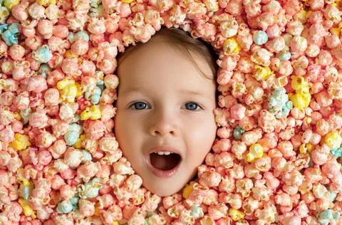 The baby's face is surrounded by a large amount of colorful sweet popcorn. Stock Photos