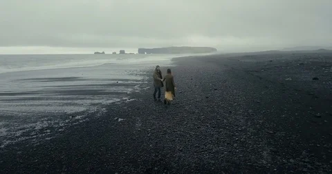Back aerial view of young hipster couple walking on the black volcanic beach in Stock Footage