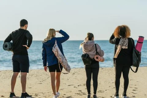Back of four people standing facing the sea while carrying yoga mats Stock Photos