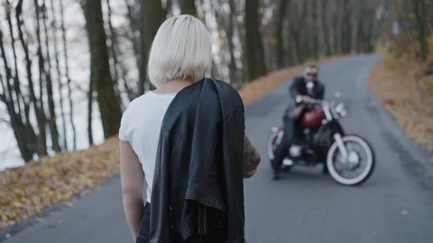 Back look of blonde comes to her smiling biker sitting at motorcycle in park Vidéo 126009791