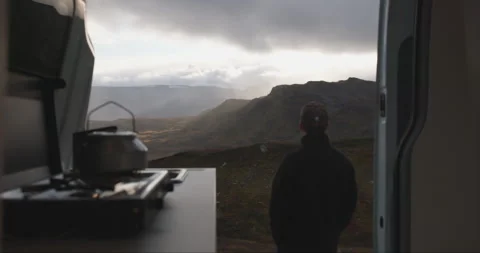 Back of male looking at glowing rainy clouds by camper van door in Norway Stock Footage 255221427