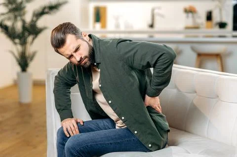 Back problems concept. Sad caucasian man, in casual wear, sit on sofa in living Stock Photos