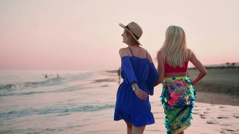 Back shoot of two stylish beautiful girls walking by cost side holding hands at Stock Footage 119385474