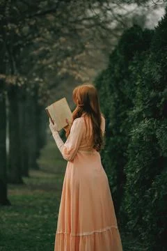 Back turned, book held aloft, she reads among trees like ghost of knowledge Stock Photos