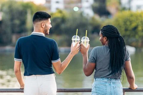 Back of two multiethnic people toasting with a shake in front of an urban river Stock Photos