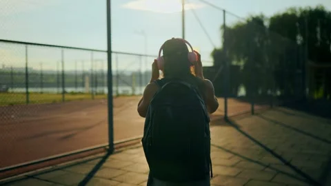 Back view of adolescent putting pink headphones on near sports fence at sunset. Vidéo 339809902
