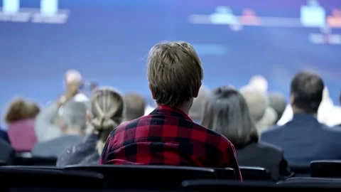 Back view of an attendee in a red plaid shirt seated among a diverse audience in Video stock 325671542
