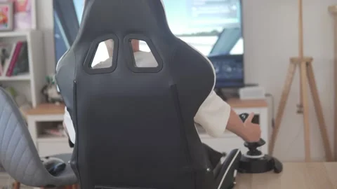 Back view of a blond boy sitting in a racing-style gaming chair while playing a Stock Footage 312339938