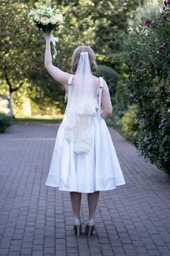 Back view of a bride in a tea-length white wedding dress and lace veil, raising  Stock Photos