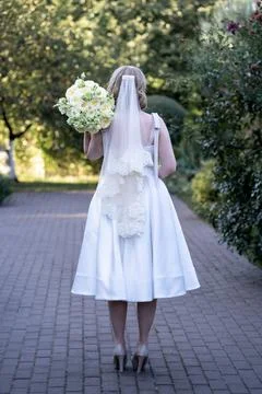 Back view of a bride in a tea-length white wedding dress and lace veil, holding  Stock Photos