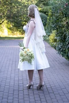 Back view of a bride in a tea-length white wedding dress and lace veil, holding  Stock Photos