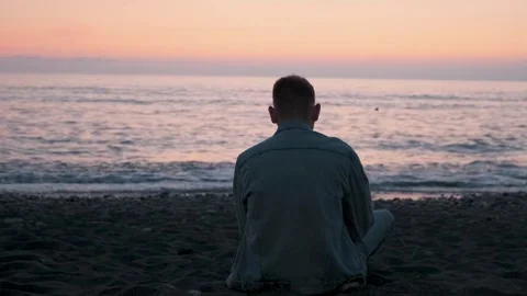Back view of Caucasian man sitting alone on black sand beach on the coast and Stock Footage 286360695