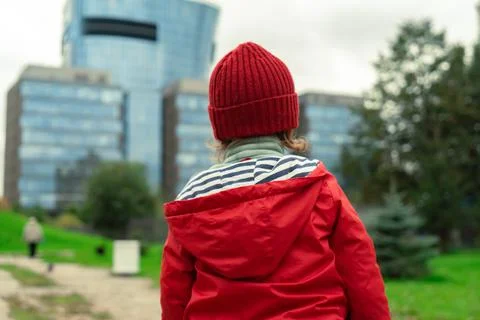 Back view of a child in a red jacket and hat standing outdoorsin the city. Stock Photos