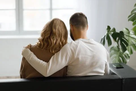 Back view of couple hugging while sitting on sofa and looking out window at home Stock Photos