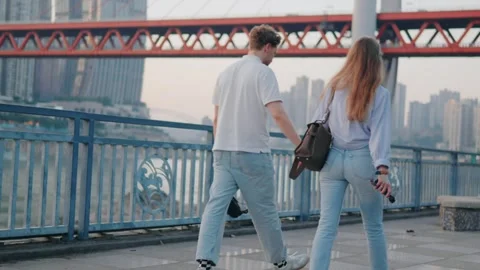 Back view of couple walking riverside promenade at sunset, urban skyline Видео 313917103