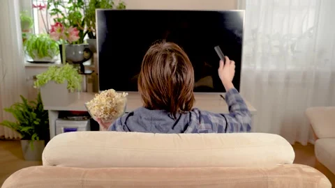 Back view of a cute young man who sits on the sofa at home and eats popcorn and Stock Footage 234869553