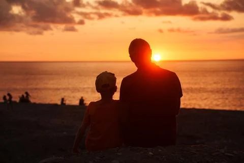 Back view of dad , son sitting at sunset on seashore enjoying summer sunset Stock Photos