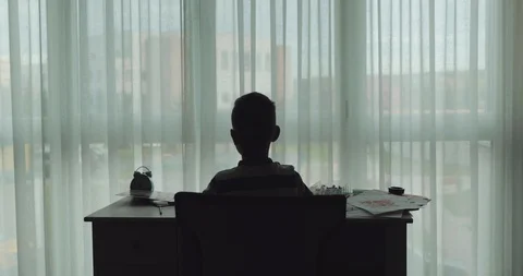 A back view of a dark silhouette of a boy sitting at the desk Stock Footage 122429613