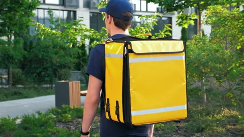Back view delivery young man courier with thermal backpack walks down street Stock Footage 157602577