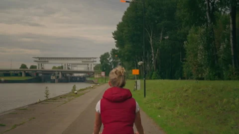 Back View Follow Shot of Blonde Woman Cycling Along Forest and River With Stock Footage 345618433