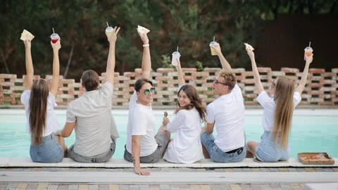 Back view of friends clinking glasses with cocktails sitting by swimming pool Stock Footage 149606362