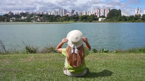 Back view of girl with hat sitting on grass in Barigui Park, Curitiba, Brazil Vídeo Stock 240142246