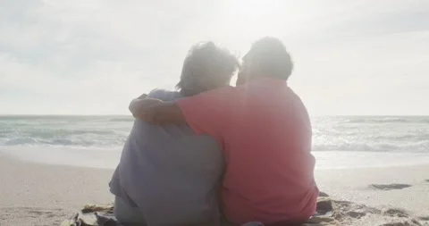 Back view of happy hispanic senior couple sitting and embracing on beach at Stock Footage 341942046